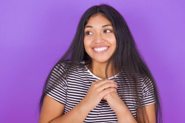 Mor arka plana karşı çizgili tişörtlü, mutlu, genç, güzel esmer kadın harika bir şey bekliyor ve yüzünün yanında el ele tutuşuyor.