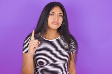 Mor arka planda çizgili tişörtlü esmer kadın işaret parmağını kaldırıyor.