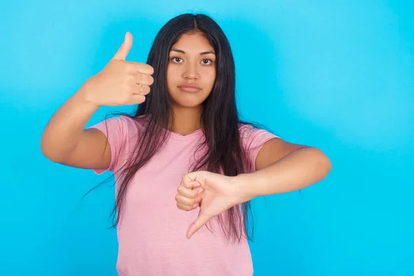 woman   showing thumb  up and thumb down, difficult choose concept. Young beautiful Hispanic brunette woman wearing pink T-shirt posing against blue background