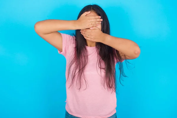 10代の少女は手で目と口を覆い ショックを受けました 感情を隠している 青い背景にポーズピンクTシャツを着ている若い美しいヒスパニックブルネット女性 — ストック写真