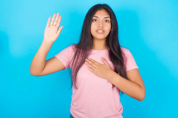 teenage girl Swearing with hand on chest and open palm, making a loyalty promise oath. Young beautiful Hispanic brunette woman wearing pink T-shirt posing against blue background