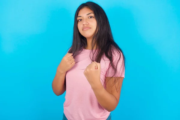 10代の少女は フェイスト防衛ジェスチャーと戦う準備ができています 怒りと怒って顔を動揺 問題を恐れ 青い背景にポーズピンクTシャツを着ている若い美しいヒスパニックブルネット女性 — ストック写真