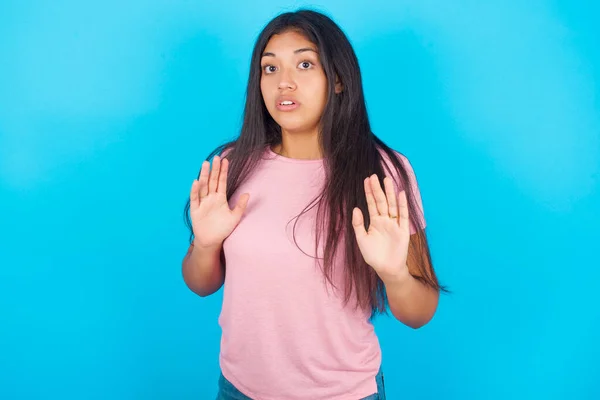 少女移开双手手掌 带着恐惧和恶心的表情表示拒绝和否认 停止和禁止 年轻美丽的西班牙裔黑发女子 身穿粉色T恤 背景为蓝色 — 图库照片