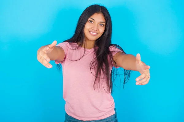 少女看着相机 张开双臂拥抱她 喜庆的表情拥抱着幸福 年轻美丽的西班牙裔黑发女子 身穿粉色T恤 背景为蓝色 — 图库照片