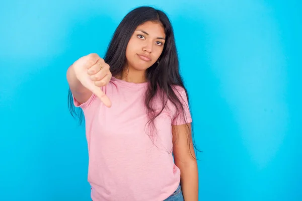 十几岁的女孩感到愤怒 失望或不高兴 带着严肃的表情垂下大拇指 年轻美丽的西班牙裔黑发女子 身穿粉色T恤 背景为蓝色 — 图库照片