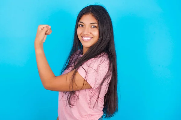强壮而快乐的女孩表现出肌肉的画像 年轻美丽的西班牙裔黑发女子 身穿粉色T恤 背景为蓝色 — 图库照片