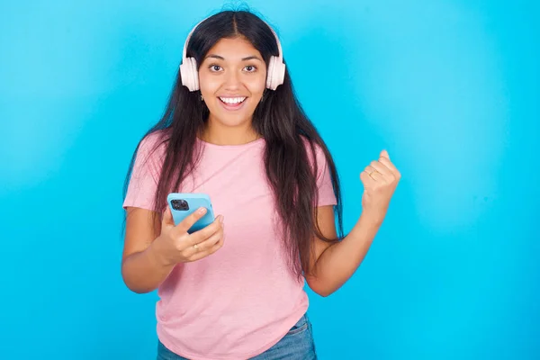 积极向上的女孩拿着与耳机相连的现代手机 从美好的情感中握紧拳头 欢呼雀跃 年轻美丽的西班牙裔黑发女子 身穿粉色T恤 背景为蓝色 — 图库照片