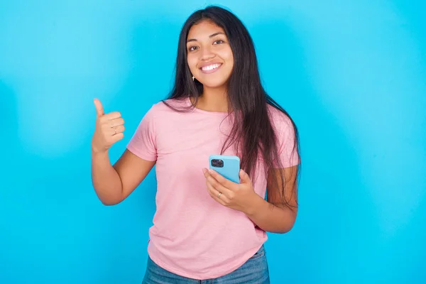 女孩使用智能手机和发短信的肖像 开心地笑着做签名大拇指 年轻美丽的西班牙裔黑发女子 身穿粉色T恤 背景为蓝色 — 图库照片