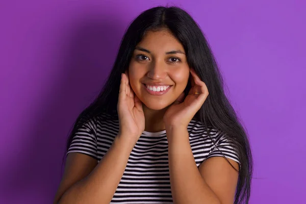 ハッピー若い美しいヒスパニックブルネット女性は指で彼女の耳を覆う紫色の背景に対してストライプTシャツ — ストック写真