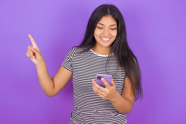 穿着条纹T恤的年轻拉丁女人微笑着 用手指着一只手拿着手机的空白处 — 图库照片