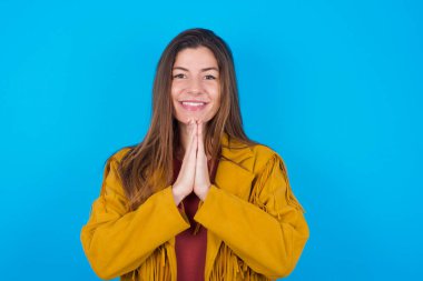  Mavi stüdyo arka planında ceket giyen kadın avuç içlerini bir arada tutuyor ve mutlu bir ifade takınıyor. Çekici bir erkek merhamet diliyor. Umutlu genç yetişkin.