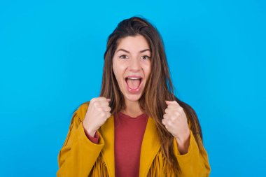  Mavi stüdyo arka planında ceketini giyen kadın başarı ve zaferle seviniyor yumruklarını zevkle sıkıyor amacına ve hedeflerine ulaştığına seviniyor. Olumlu duygular, duygular.