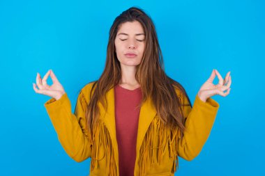 Mavi stüdyo arka planında ceket giyen genç güzel bir kadın yoga yapıyor, gözlerini kapalı tutuyor, Mudra jestinde parmaklarını tutuyor. Meditasyon, din ve ruhani uygulamalar.