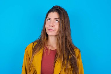 Mavi stüdyo arka planında ceket giyen genç ve güzel bir kadın endişeli bir yüze sahip dudaklarının arasından bakarak, üzgün bir şekilde önemli bir şeyi düşünüyor, ellerini aşağıda tutuyor..