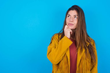 Mavi stüdyo arka planında ceket giyen düşünceli genç bir kadının portresi elini çenesinin altında tutuyor, bir şey hatırlamaya çalışıyor ya da ilgi çekici bir şey dinliyor. Gençlik kavramı.