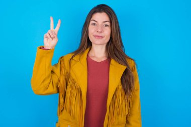 Mavi stüdyo arka planında ceketi olan genç ve güzel bir kadın iki numaralı parmaklarıyla gülümseyerek kendinden emin ve mutlu bir şekilde işaret ediyor..