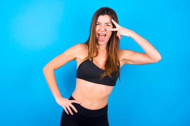 Güzel esmer kadın spor giysisi giyiyor ve gözlerinin yanında V işareti yapıyor. Boş zaman, cilveleşme, kutlama ve flört konsepti.