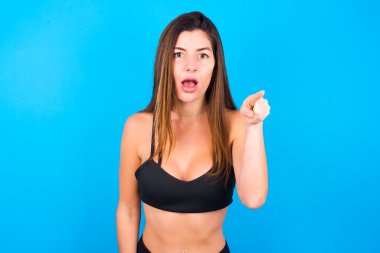 Şok olmuş güzel esmer kadın spor giysileri giyiyor ve kameraya işaret parmağıyla bakıyor. Sürpriz ve reklam konsepti.