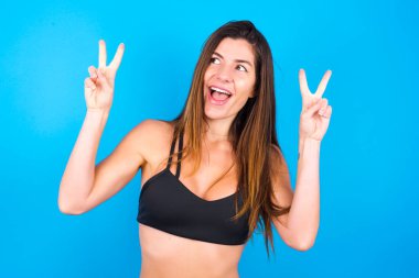 Spor giysili, neşeli, esmer bir kadının yalnız pozu iki elinle barış ya da zafer işareti yapar..