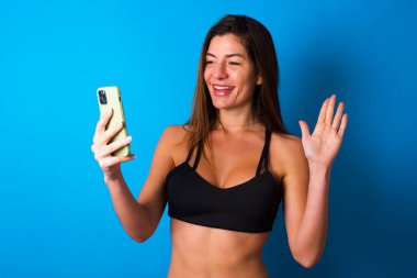 Mutlu esmer kadın portresi spor kıyafetleri giymiş selfie çekiyor ve el sallıyor, video görüşmesinde iletişim kuruyor, internette sohbet ediyor..