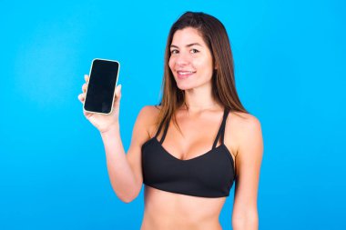 Gülümseyen esmer kadın spor giysisi giyiyor ve boş telefon ekranı gösteriyor. Reklam ve iletişim konsepti.