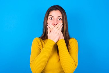 Sarı balıkçı yaka kazak giyen, ağzı iki eliyle örten afallamış genç bir kadın bir şeyden ya da çarpıcı dedikodular duyduktan sonra korkuyor..