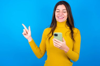 Mavi stüdyo arka planında sarı süveter giyen gülümseyen güzel bir kadın bir elinde telefonu tutan boş bir yeri işaret ediyor.
