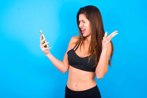 Photo of outraged annoyed  brunette woman wearing sportswear holds cell phone, makes call, argues with colleague,  expresses negative emotions. People and anger.