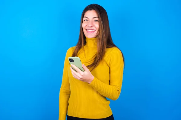 年轻而漂亮的女人 身穿黄色毛衣 头戴蓝色的工作室背景 手握手机 喜庆好消息 用现代手机 — 图库照片