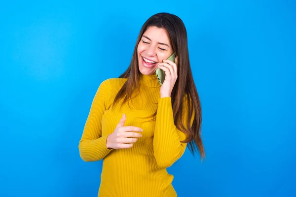 青いスタジオの背景に黄色いセーターを着ているおかしい若い美しい女性は幸せに笑い 友人によって楽しまれて 目を閉じる電話会話を持っています — ストック写真