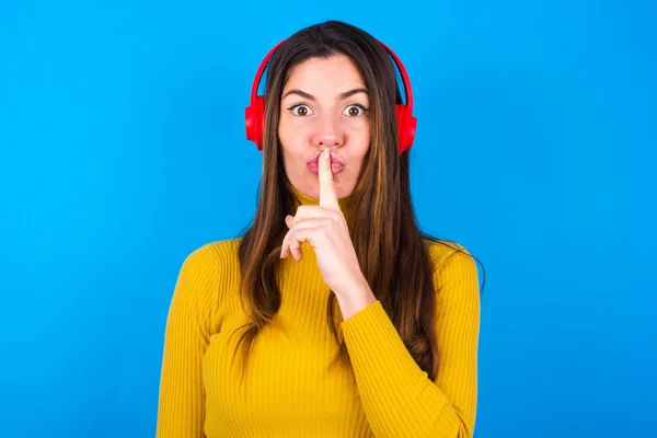woman wearing turtleneck sweater making hush gesture with finger on her lips wearing  wireless headphones. Be quiet.