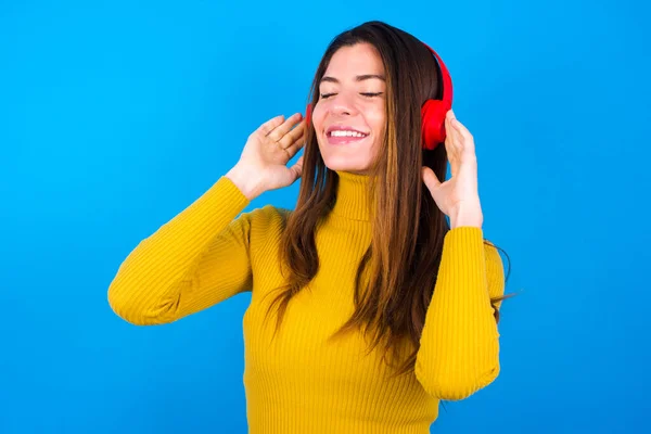 woman wearing turtleneck sweater with headphones on head, listens to music, enjoying favourite song with closed eyes, holding hands on headset.