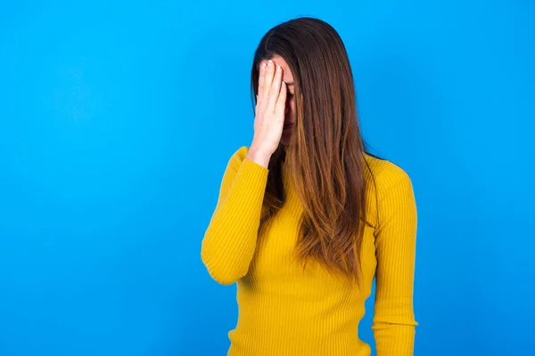 青いスタジオの背景に黄色いセーターを着た若い美しい女性は 泣きながら手で顔を覆う悲しい表情をしています うつ病コンセプト — ストック写真