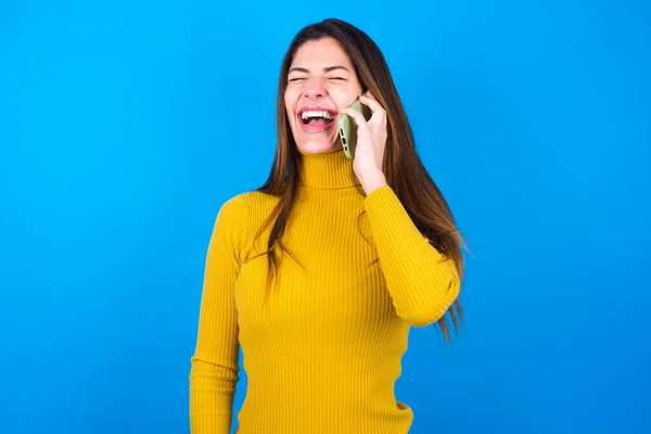 青いスタジオの背景に黄色いセーターを着ている幸せな若い美しい女性を過度に発言 電話会話中に友人からの面白い話を肯定的に聞きます — ストック写真