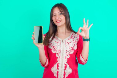 Genç, güzel, beyaz bir kadın elinde akıllı telefon tutuyor ve iyi olduğunu gösteriyor.