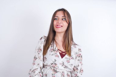 Beyaz arka planda çiçek desenli elbiseler giyen genç ve güzel bir kadın stüdyoda aptal bir yüz ifadesi takınıyor.