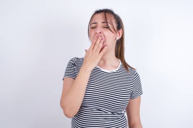 Beyaz arka plan üzerine çizgili tişört giyen genç bir Arap kadın stüdyoda esniyor.