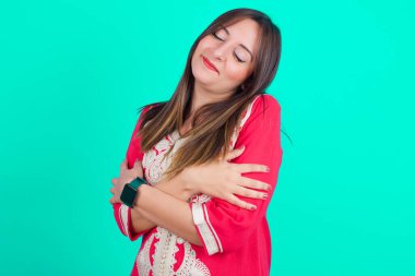 Genç ve güzel Faslı bir kadın geleneksel kaftan elbiseler giyip yeşil arka planda kendine sarılıyor mutlu ve pozitif, kendinden emin gülümsüyor. Sevgi ve özgüven.