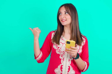 Genç, güzel, beyaz bir kadın akıllı telefon kullanıyor. Baş parmağıyla işaret ediyor ve gülümsüyor.