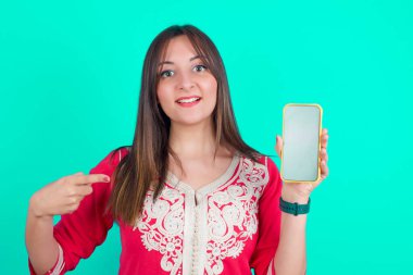 Çekici, neşeli, beyaz tenli, güzel bir kadın, elinde telefonla siyah ekranı gösteriyor.