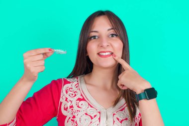 Genç, güzel, beyaz bir kadın görünmez bir hizalama aleti tutuyor ve mükemmel düz dişlerine işaret ediyor. Diş sağlığı ve güven kavramı.