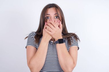 Beyaz arka plan üzerine çizgili tişört giyen afallamış genç bir Arap kadın iki elini de ağız üstüne koyup hayret verici bir şeyden korkuyor.