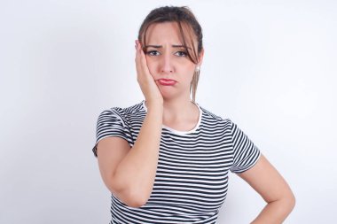 Beyaz arka plan üzerine çizgili tişört giyen üzgün, yalnız bir Arap kadın yanağına dokunuyor alt dudağını ısırıyor ve hoşnutsuzca bakıyor. Kötü duygular