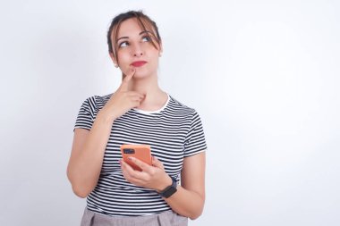 Tişört giyen güzel beyaz kadın bir şey hakkında derin düşünüyor, modern cep telefonu kullanıyor, iyi bir mesaj uydurmaya çalışıyor, işaret parmağını dudaklarının yakınında tutuyor..