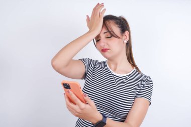 Üzgün, üzgün, beyaz tişört giyen genç bir kadın, unutulan bir şeyi yüzüne vuruyor. Cep telefonu tutmak, üzüntü ve pişmanlık ifade ediyor.