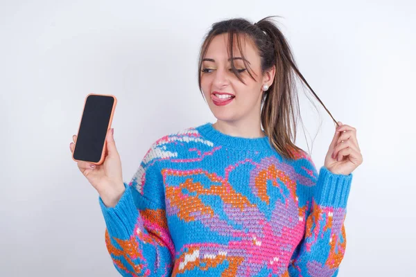 照片上漂亮的阿拉伯年轻女子穿着色彩艳丽的针织毛衣 背景洁白 展示手机屏幕 手挽着发尾 — 图库照片