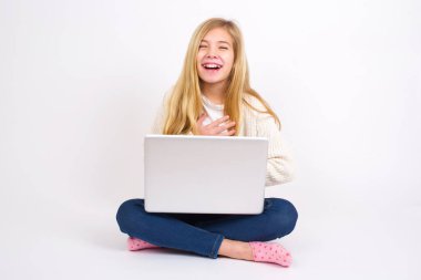 Beyaz arka planda Lotus pozisyonunda oturan beyaz tenli genç kız ellerini çapraz tutar, şakaya yüksek sesle güler, günlük kıyafetler giyer..