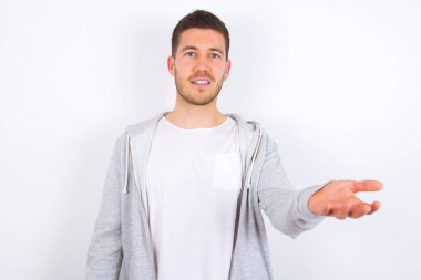 Beyaz arka plan üzerine günlük kıyafetler giymiş genç beyaz bir adam gülümseyerek dostça bir şey sunuyor. Açık elle veya el sıkışarak selamlama ve hoş geldin. Başarılı bir iş..