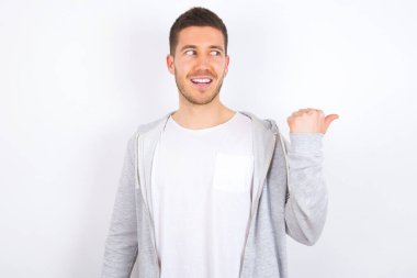 Şaşırmış, beyaz arka plan üzerine günlük kıyafetler giyen, ağzı ve gözlerini geniş bir şekilde açan, baş parmağıyla işaret eden genç, beyaz bir adam, garip bir şey gösteriyor. Reklam kavramı.
