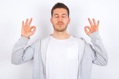 Beyaz arka plan üzerine günlük kıyafetler giyen genç beyaz bir adam yoga yapıyor, gözlerini kapalı tutuyor, Mudra jesti için parmaklarını tutuyor. Meditasyon, din ve ruhani uygulamalar.
